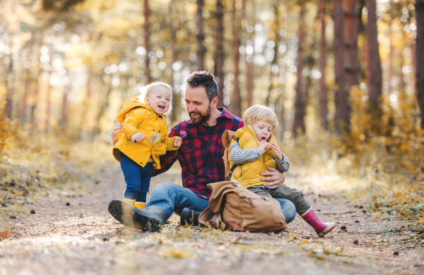 A mature father with toddler children sitting on the ground in an autumn forest, having fun.