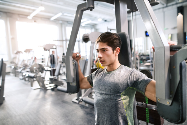 Young hispanic fitness man in gym working on butterfly fitness machine with weights, flexing muscles