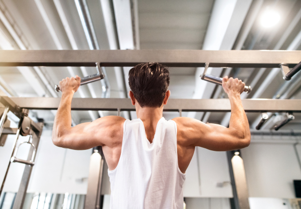 Young hispanic fitness man in gym working out, doing pull-ups on horizontal bar. Rear view.