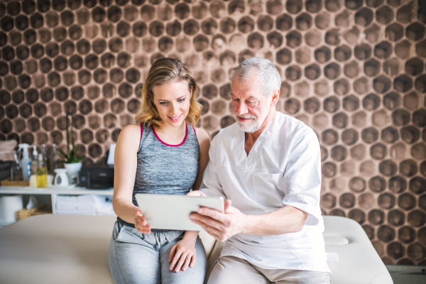 Senior male physiotherapist and young woman with tablet sitting on bed.