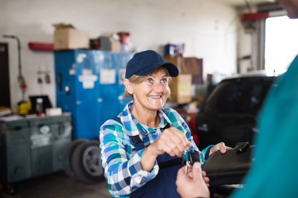 Female mechanic repairing a car. A senior woman giving a key to an unrecognizable customer.