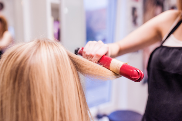 Hands of unrecognizable professional hairdresser creating hairstyle for her beautiful blond client. Straightening iron, blonde female customer.