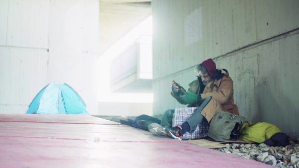 A homeless beggar man sitting on a ground outdoors in city, taking something out of a bag.