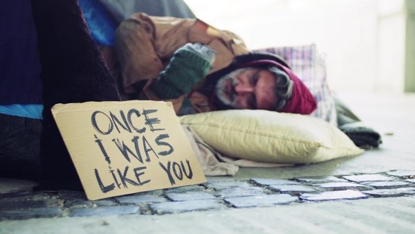 Homeless beggar man lying outdoors in city in a sleeping bag, cardboard sign near him.