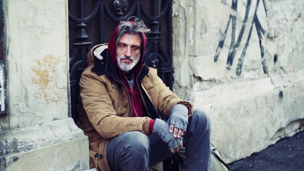 A homeless beggar man sitting in front of gate outdoors in city asking for money donation.