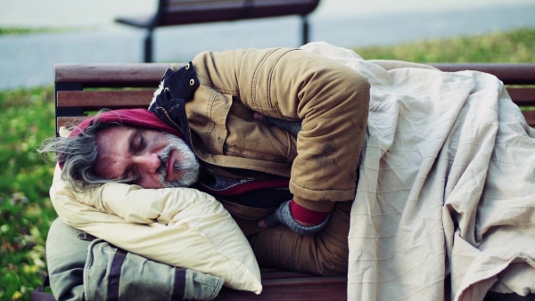 A homeless beggar man with a pillow lying on bench outdoors in city, sleeping.