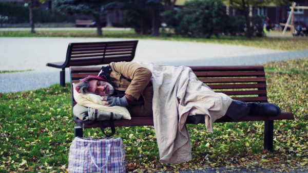 A homeless beggar man with a pillow lying on bench outdoors in city, sleeping.