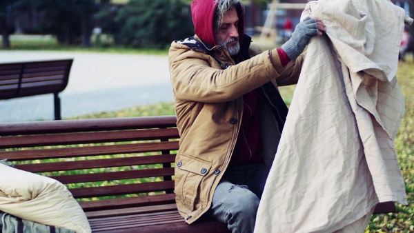 A homeless beggar man with a pillow lying down on bench outdoors in city.