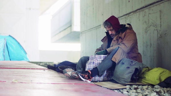 A homeless beggar man sitting on a ground outdoors in city, looking for something in a bag.