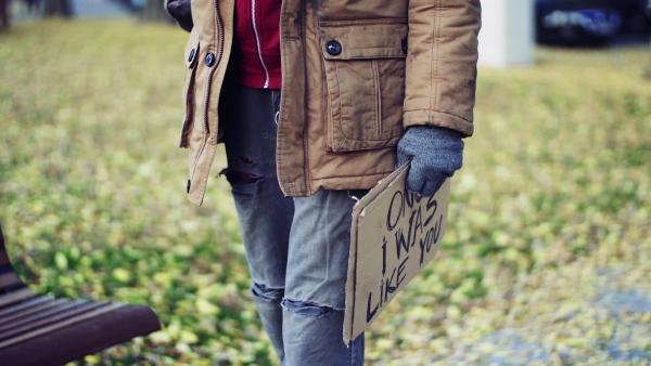 Midsection of homeless beggar man walking outdoors in park, holding a cardboard sign.