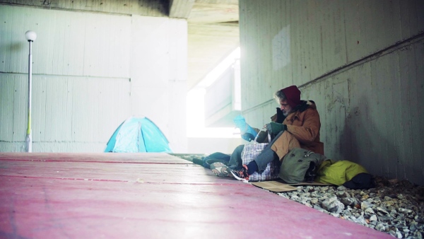 A homeless beggar man sitting on a ground outdoors in city, putting things in a bag. Slow motion.