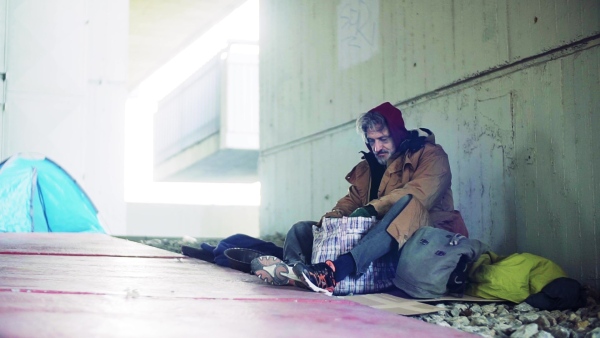 A homeless beggar man sitting on a ground outdoors in city, looking for something in a bag. Slow motion.