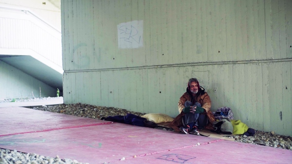 A homeless beggar man sitting on a ground outdoors in city, leaning on a wall. Slow motion.