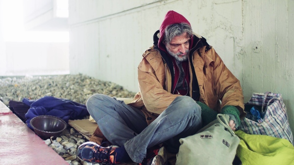 A homeless beggar man sitting on a ground outdoors in city, looking for something in a bag.