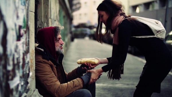 A young and kind woman giving food to homeless beggar man sitting outdoors in city. Slow motion.