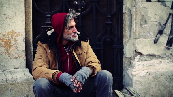 A homeless beggar man sitting in front of gate outdoors in city asking for money donation. Slow motion.