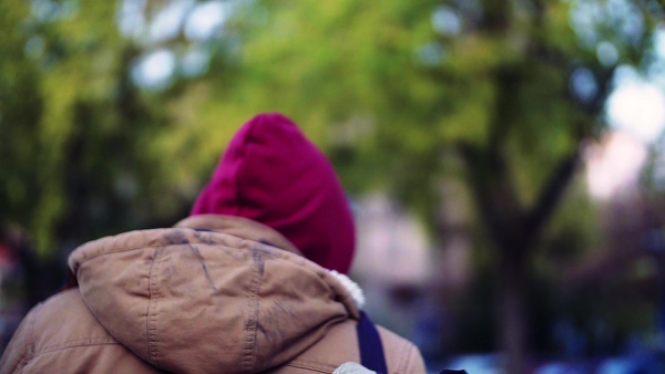 A rear view of homeless beggar man with hood on head walking outdoors in city, looking back. Slow motion.