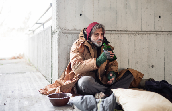 A front view of homeless beggar man sitting outdoors in city asking for money donation, holding a bottle of alcohol. Copy space.