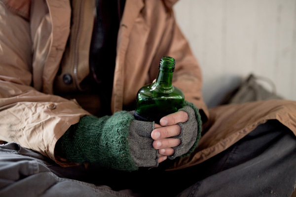 A midsection view of homeless beggar man sitting outdoors in city asking for money donation, holding a bottle of alcohol.