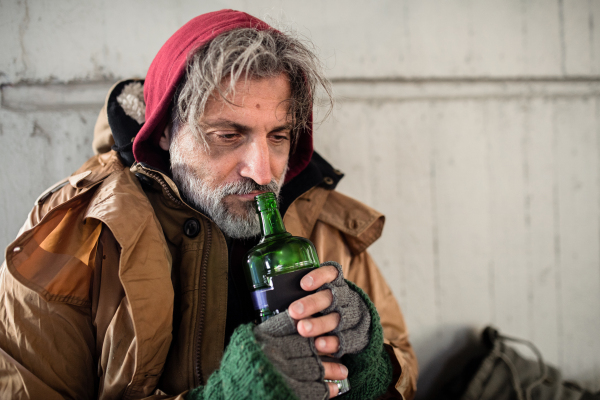 A front view of homeless beggar man sitting outdoors in city asking for money donation, holding a bottle of alcohol. Copy space.