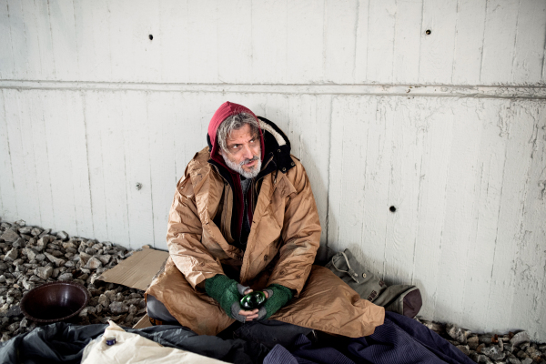 A front view of homeless beggar man sitting outdoors in city asking for money donation, holding a bottle of alcohol. Copy space.