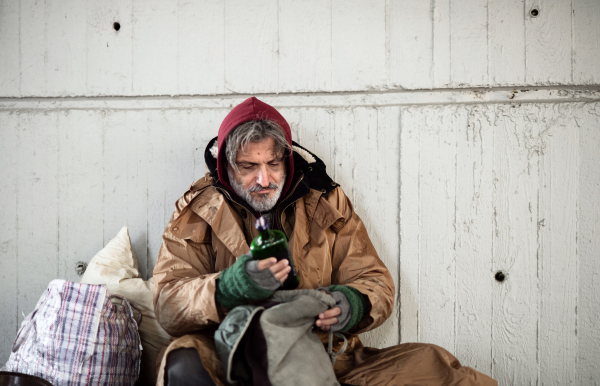 A front view of homeless beggar man sitting outdoors in city asking for money donation, holding a bottle of alcohol. Copy space.