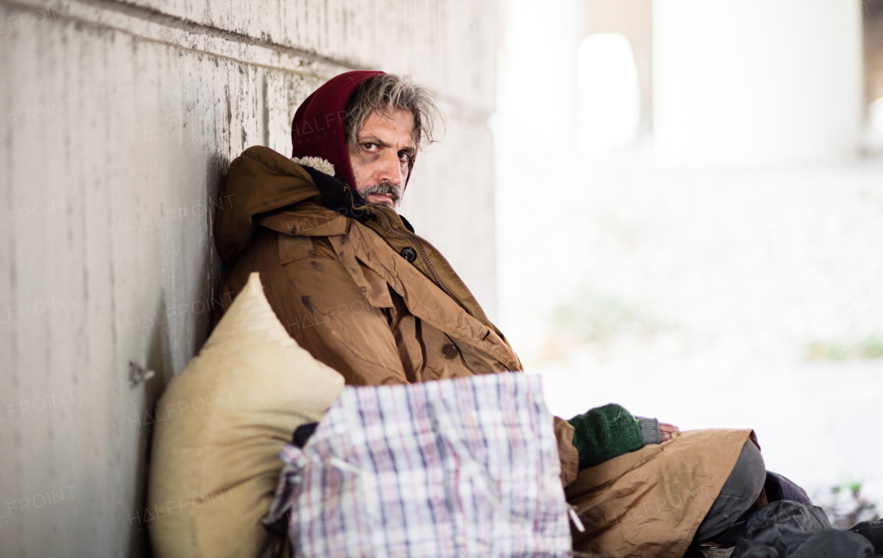 A side view of serious homeless beggar man sitting outdoors, leaning against a wall. Copy space.