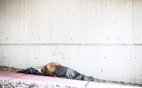 A rear view of homeless beggar man lying on the ground outdoors in city asking for money donation, sleeping. Copy space.