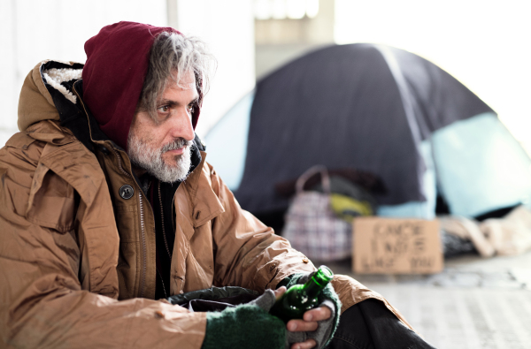 A homeless beggar man sitting outdoors in city asking for money donation, holding a bottle of alcohol. Copy space.