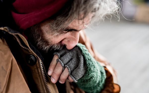 A close-up view of homeless beggar man standing outdoors in city, rubbing nose.