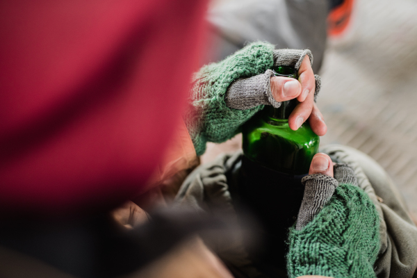 A midsection view of homeless beggar man sitting outdoors in city asking for money donation, holding a bottle of alcohol. A close-up.