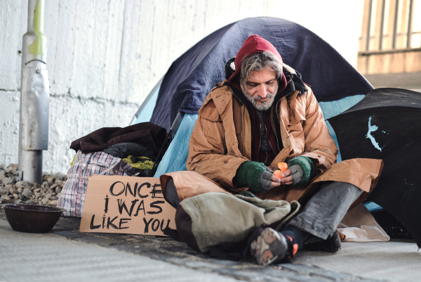 A homeless beggar man with a carboard sign sitting outdoors in city asking for money donation.