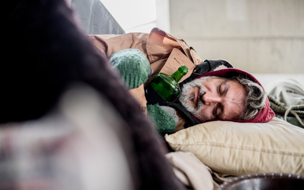 A homeless beggar man lying on the ground outdoors in city asking for money donation, holding bottle and sleeping.