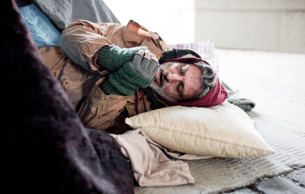 A homeless beggar man lying on the ground outdoors in city asking for money donation, warming up hands.