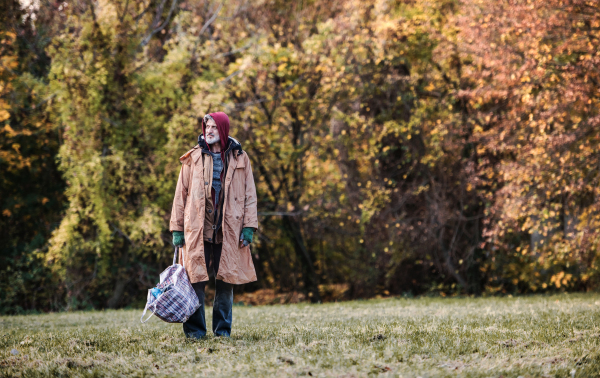 Homeless beggar man standing outdoors in park in autumn, holding bag. Copy space.