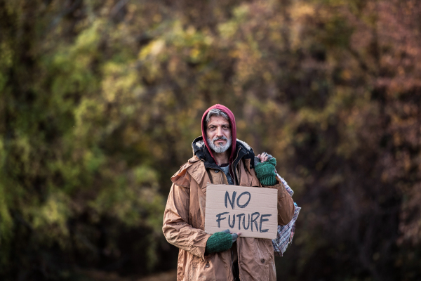 A portrait of homeless beggar man standing outdoors in park in autumn, holding bag and no future cardboard sign. Copy space.