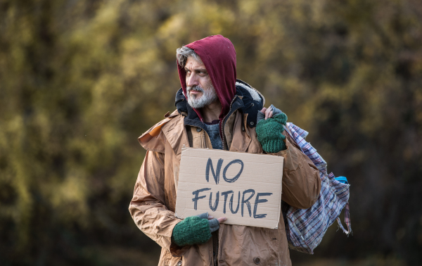 A portrait of homeless beggar man standing outdoors in park in autumn, holding bag and no future cardboard sign. Copy space.