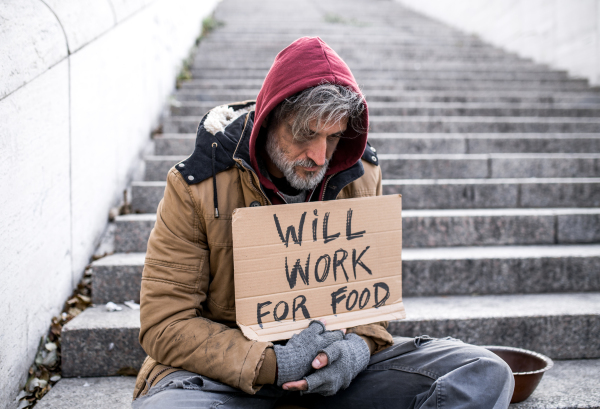 A homeless beggar man sitting on staircase outdoors in city asking for money donation.