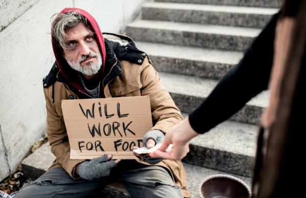 An unrecognizable woman giving money to homeless beggar man sitting in city.