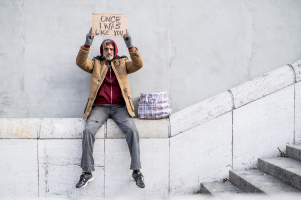 Homeless beggar man sitting outdoors in city holding will work for food cardboard sign above his head, asking for money donation. Copy space.