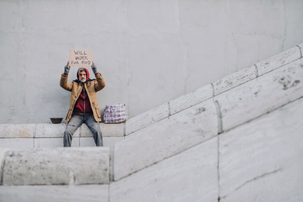 Homeless beggar man sitting outdoors in city holding will work for food cardboard sign, asking for money donation. Copy space.