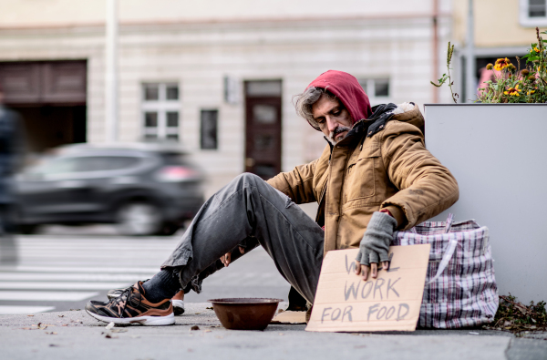 A homeless beggar man with a carboard sign sitting outdoors in city asking for money donation.