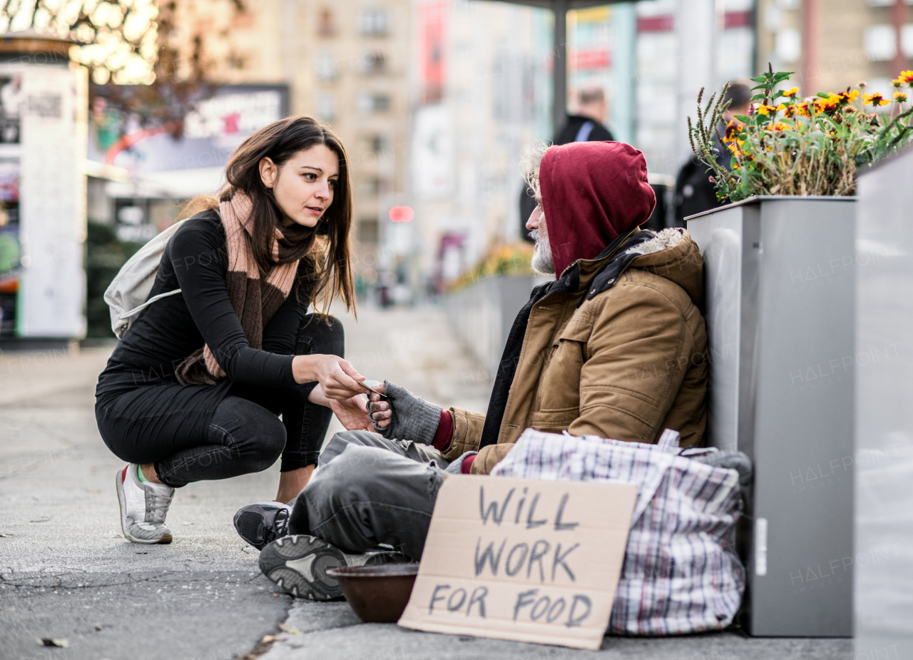 A young woman giving money to homeless beggar man sitting outdoors in city.