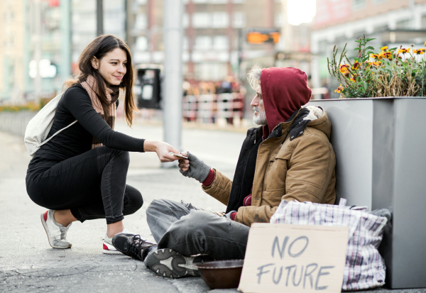 A young woman giving money to homeless beggar man sitting outdoors in city.