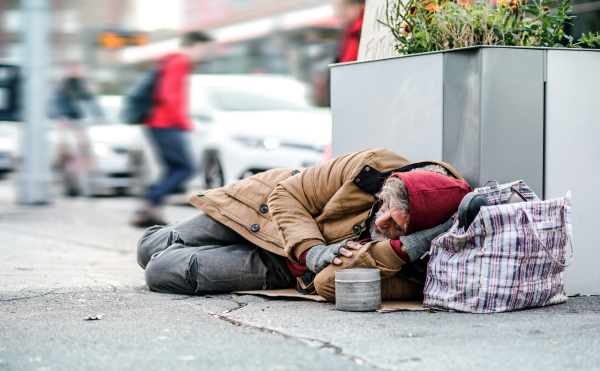 A homeless beggar man lying on the ground outdoors in city asking for money donation.