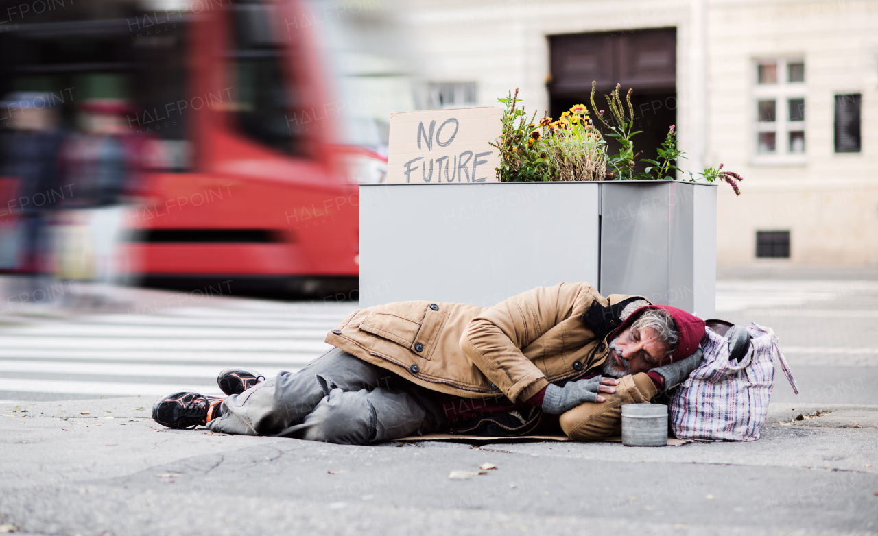 A homeless beggar man lying on the ground outdoors in city asking for money donation.