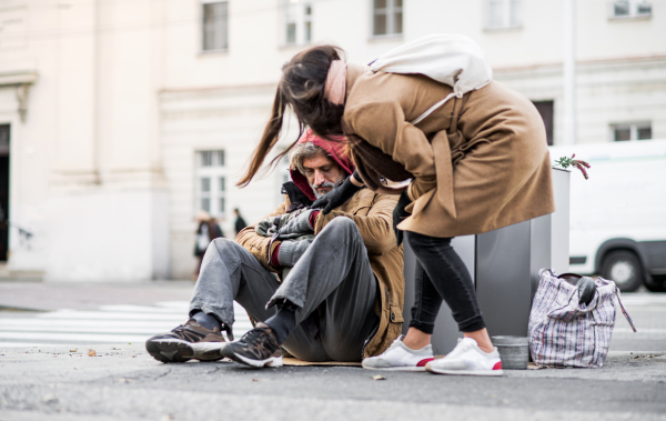 A young woman giving money to homeless beggar man sitting outdoors in city.