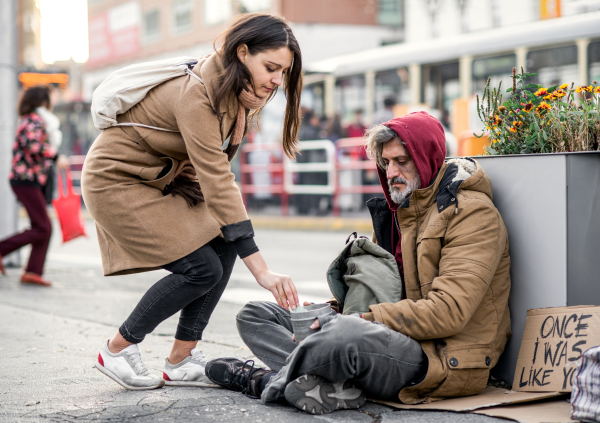 A young woman giving money to homeless beggar man sitting outdoors in city.