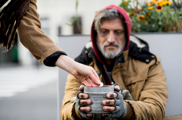 Unrecognizable woman giving money to homeless beggar man sitting outdoors in city.