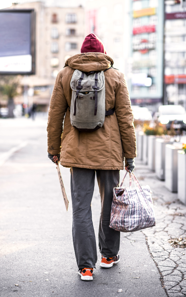 A rear view of homeless beggar man with backpack walking outdoors in city, holding bag.